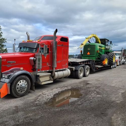 Red semi truck hauling yellow and green agricultural harvester on flatbed trailer