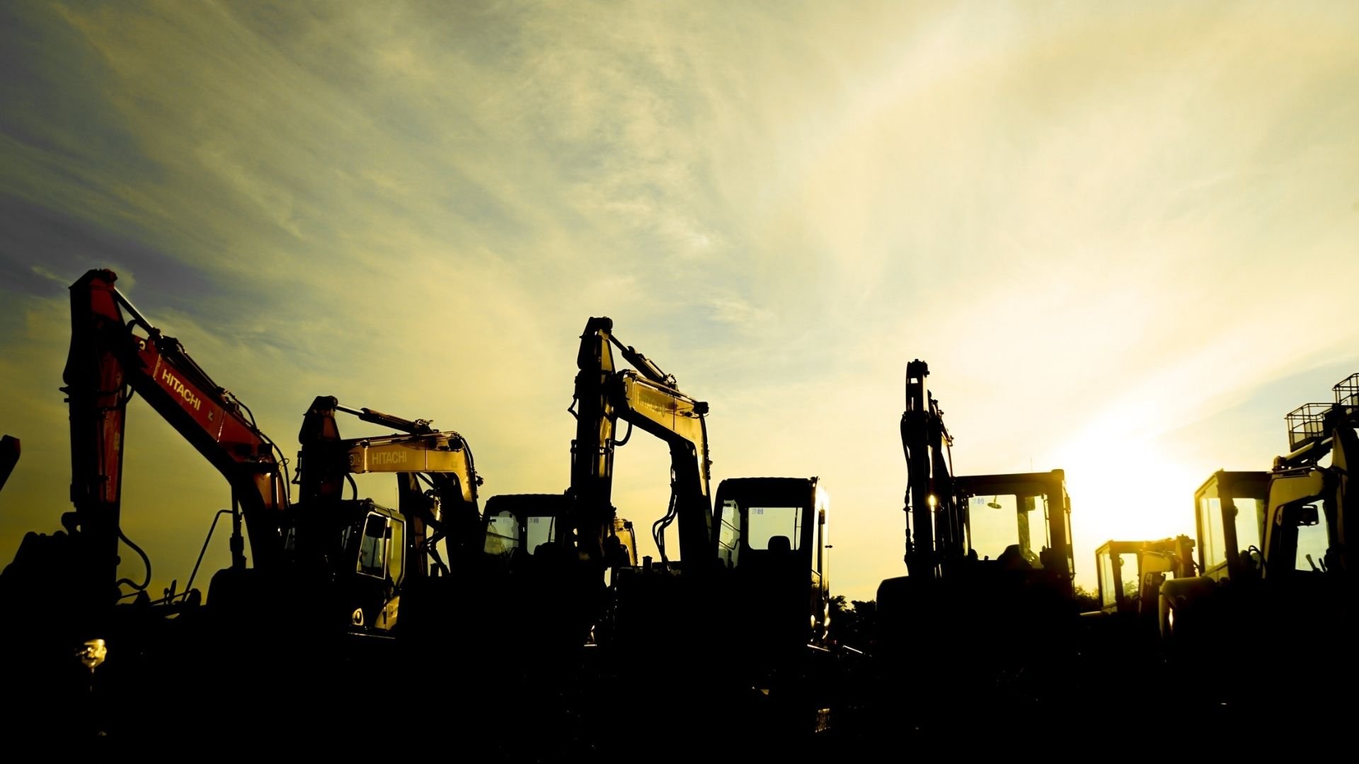 Row of silhouetted excavators and heavy machinery at sunset or sunrise.