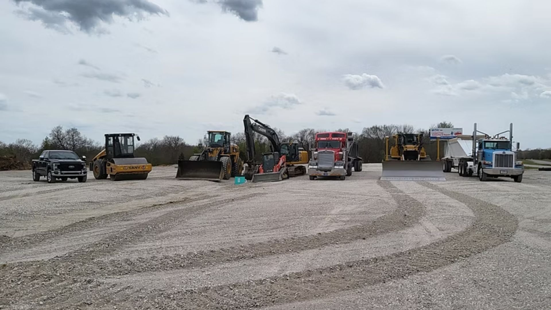 Heavy construction equipment and vehicles lined up on a gravel yard under cloudy sky.